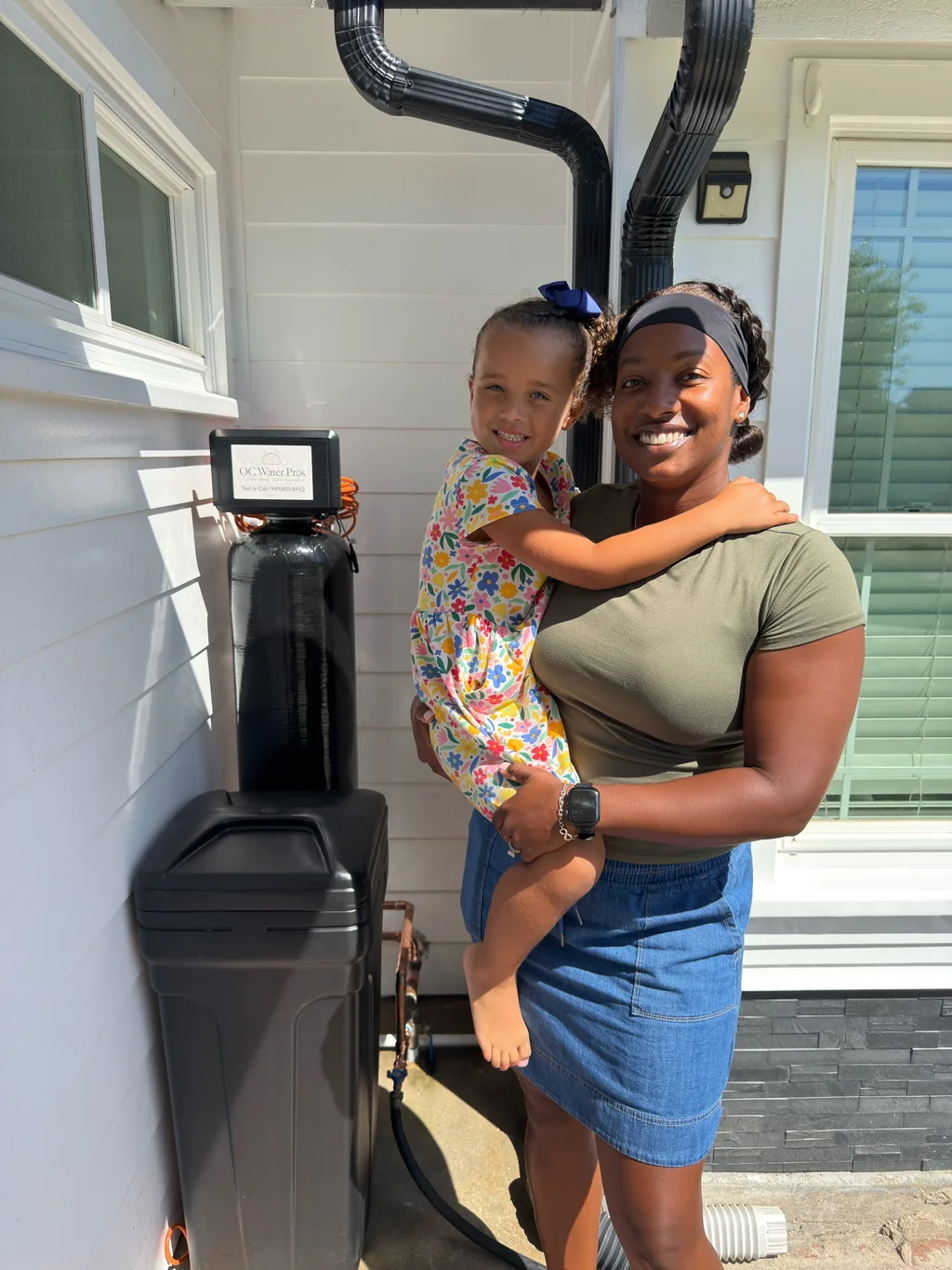 A smiling homeowner holding her young daughter next to a newly installed softener and brine tank
