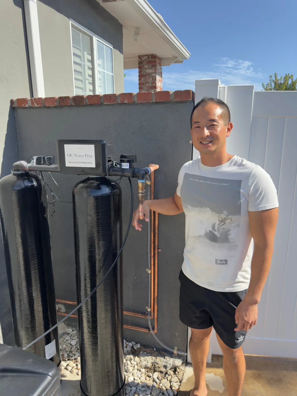 A homeowner standing beside two black whole-home filtration tanks mounted against a side-yard wall